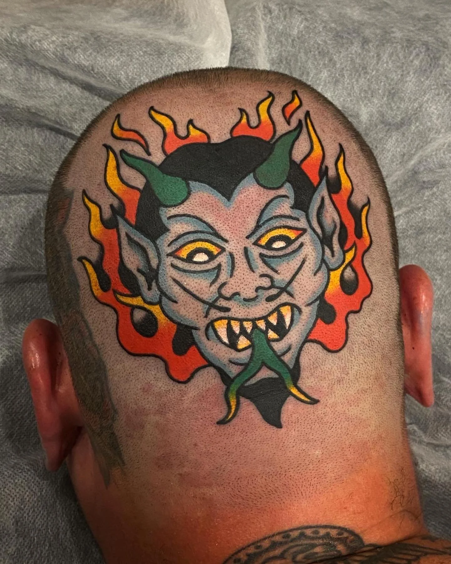 Tattoo of a sinister, cartoonish demon face with horns, pointed ears, yellow eyes, sharp teeth, and a long tongue, surrounded by flames.
