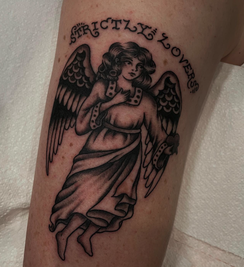 Tattoo of an angel with detailed wings, wearing a flowing gown, with a hand over chest, and the words "STRICTLY LOVERS" curved above.