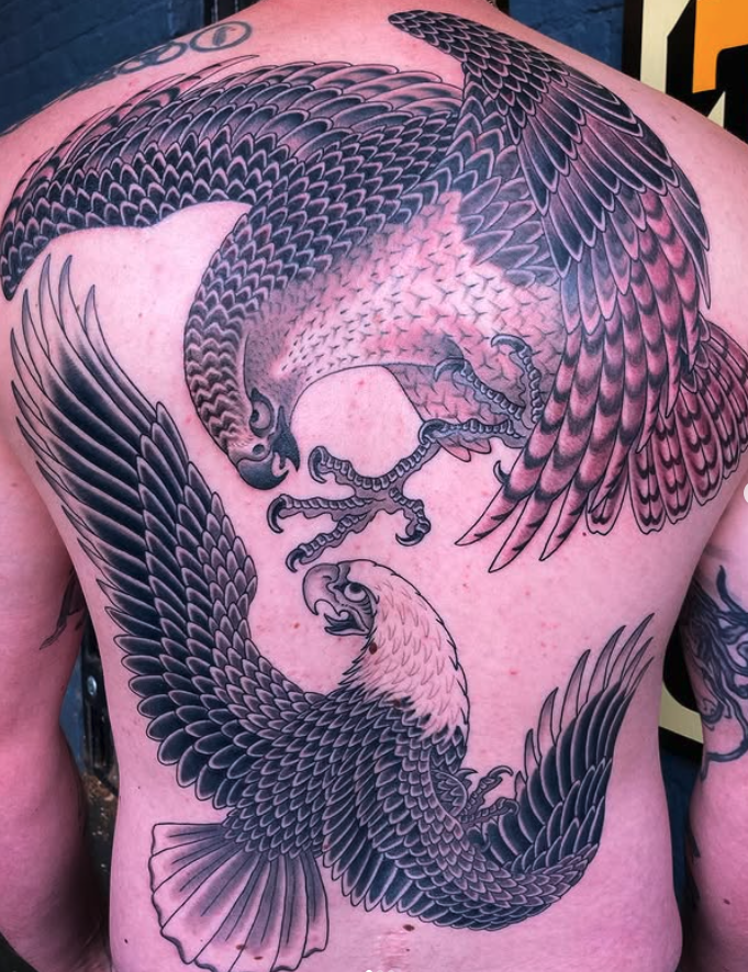 A detailed black and gray tattoo of two eagles in mid-flight on a person's back, with wings spread wide and talons extended.