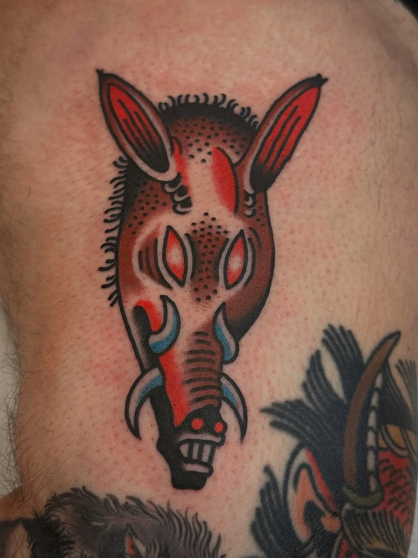 Tattoo of a stylized animal face, possibly a wolf or fox, with pointed ears, red eyes, and a long snout, in black, red, and blue ink.