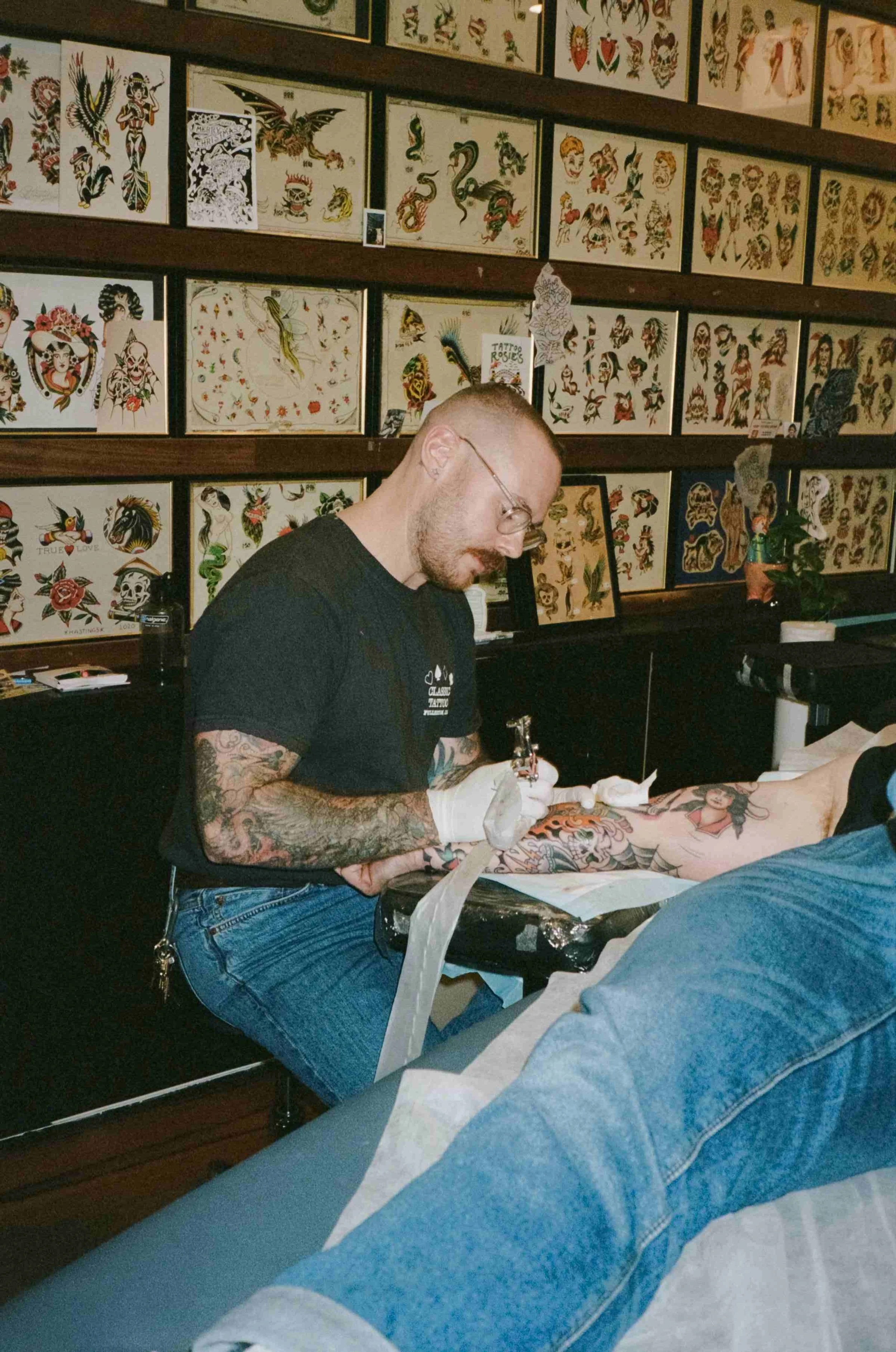 A tattoo artist working on a client's arm in a tattoo shop decorated with tattoo flash art on the walls.