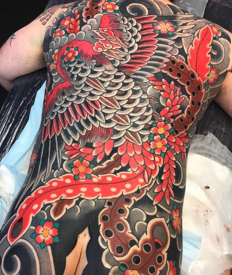 Colorful Japanese-style tattoo on person's back featuring a phoenix, flowers, and swirling cloud designs.
