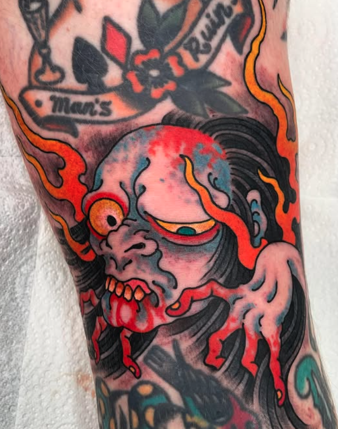 Tattoo of a cartoonish, zombie-like creature with a large head, one eye, exposed teeth, and tongue hanging out, surrounded by flaming details and dark flowing hair.