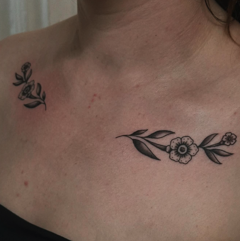 A person's upper chest and shoulder with two black ink floral tattoos, one on the shoulder and one on the chest, featuring leaves and flowers.