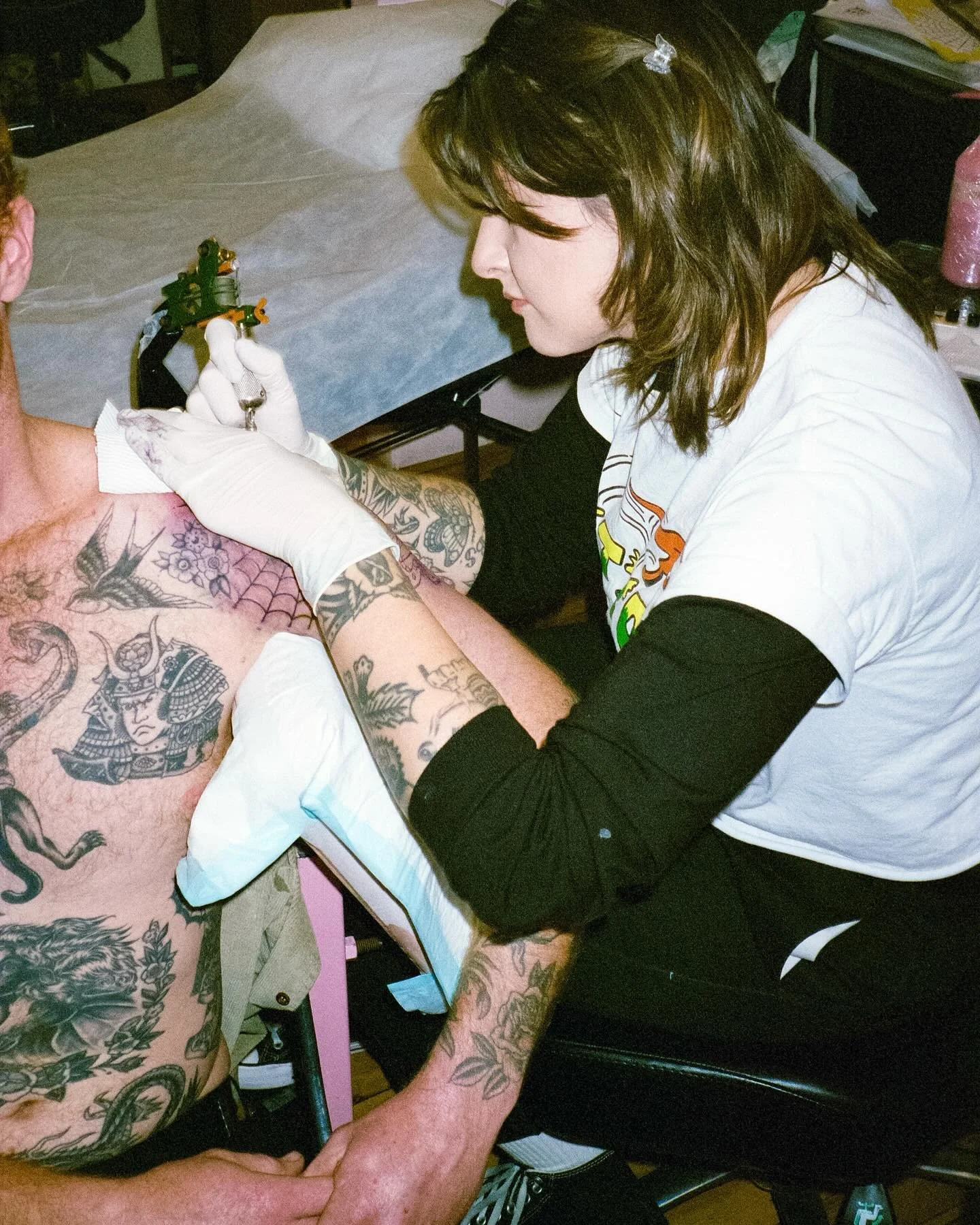 A woman is tattooing a man's chest and arm at a tattoo parlor, with tattoo equipment and inks visible on a nearby table.