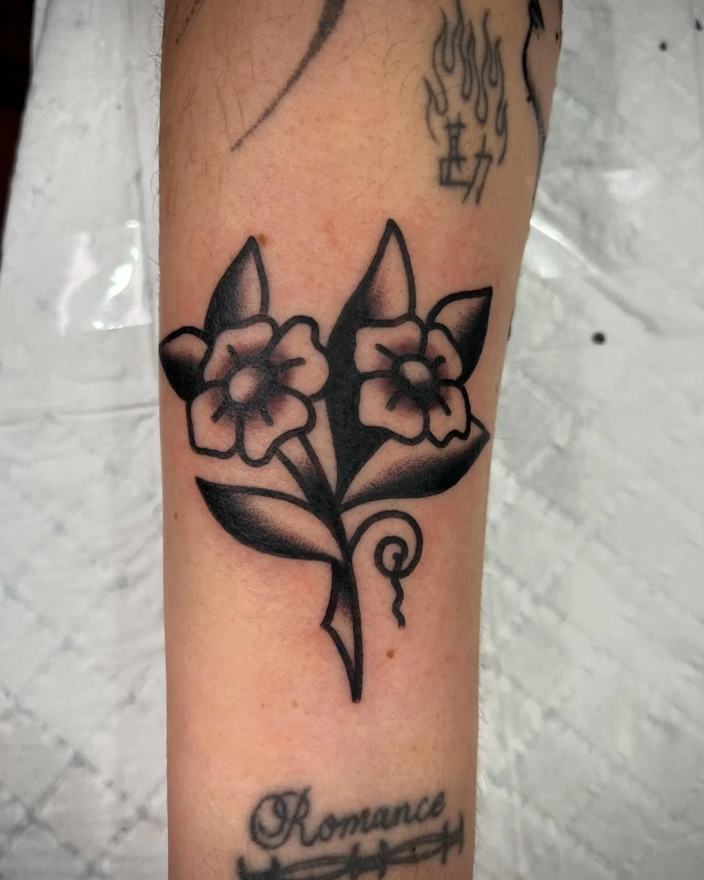 Black outline tattoo of a branch with two flowers and leaves on person's forearm.
