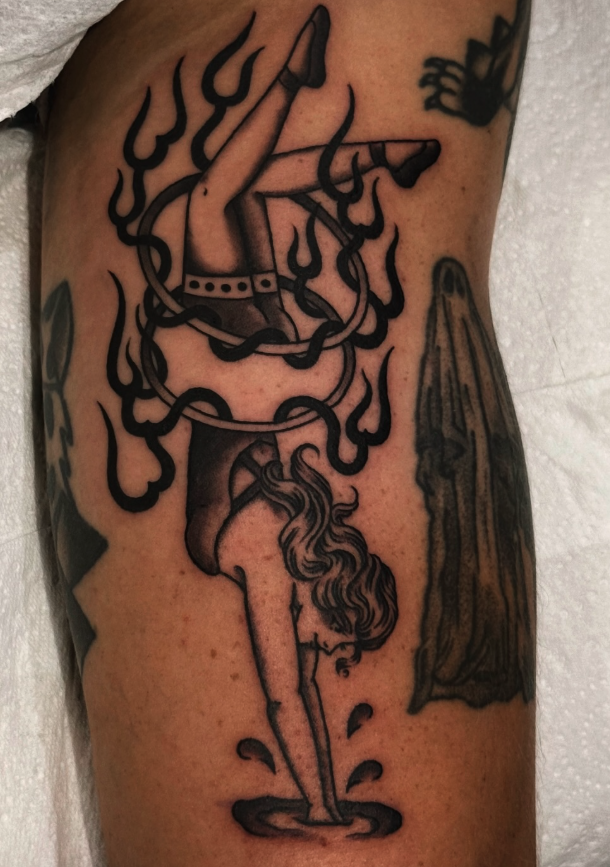 Tattoo of a woman with long hair, bending over with her hands in water, surrounded by flames, on a person's arm.