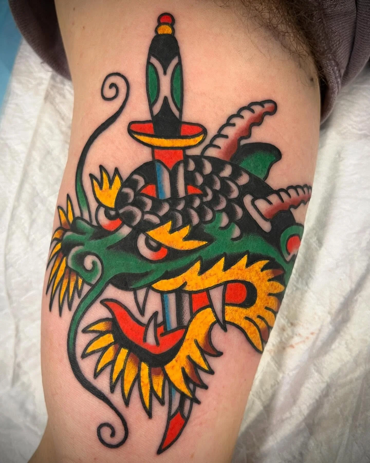 Colorful tattoo of a dragon with a sword in its mouth, featuring vivid green, yellow, orange, red, and black outline details.