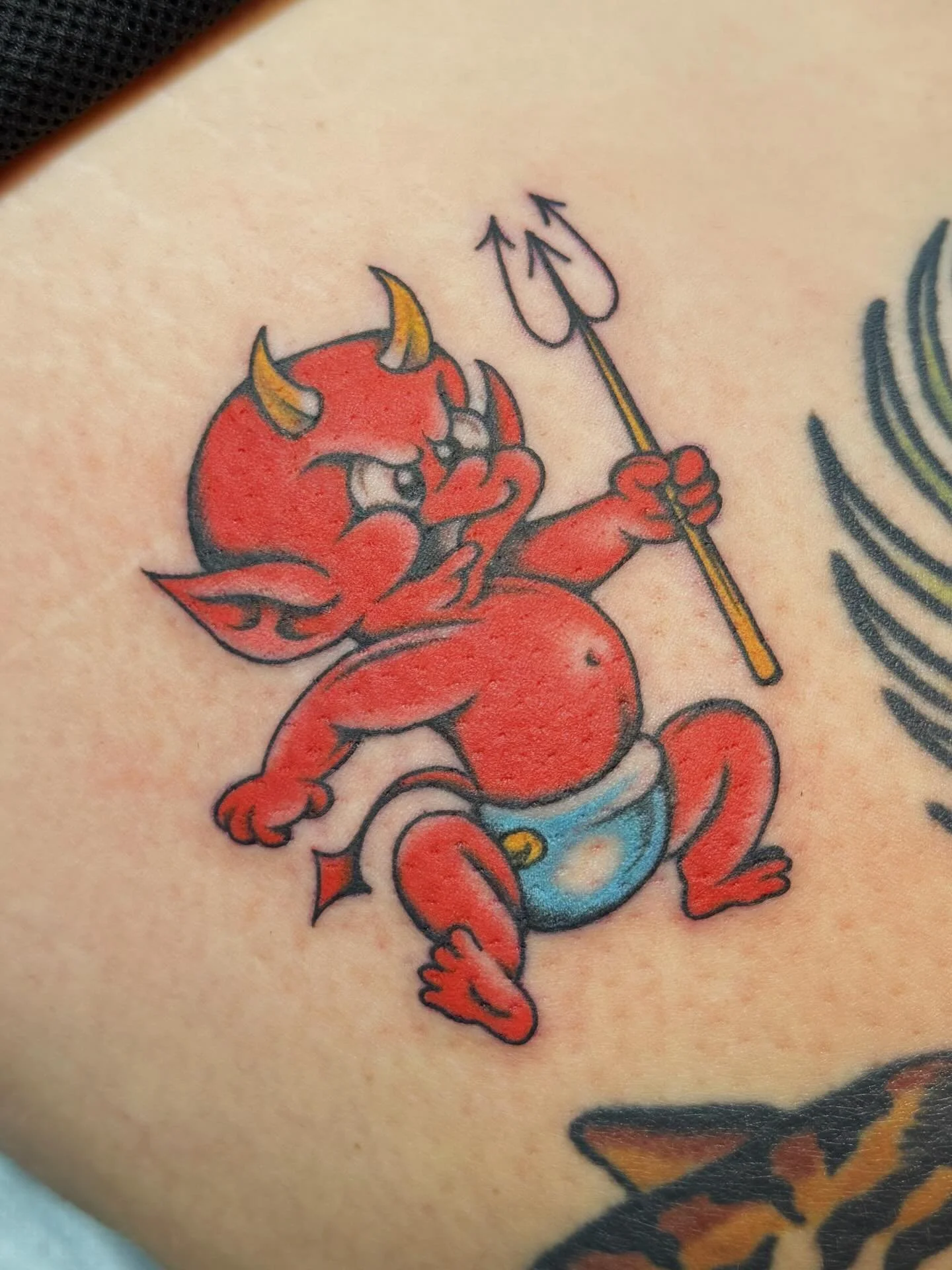 Tattoo of a red devil with horns, holding a trident, with a diaper, in a cartoon style.