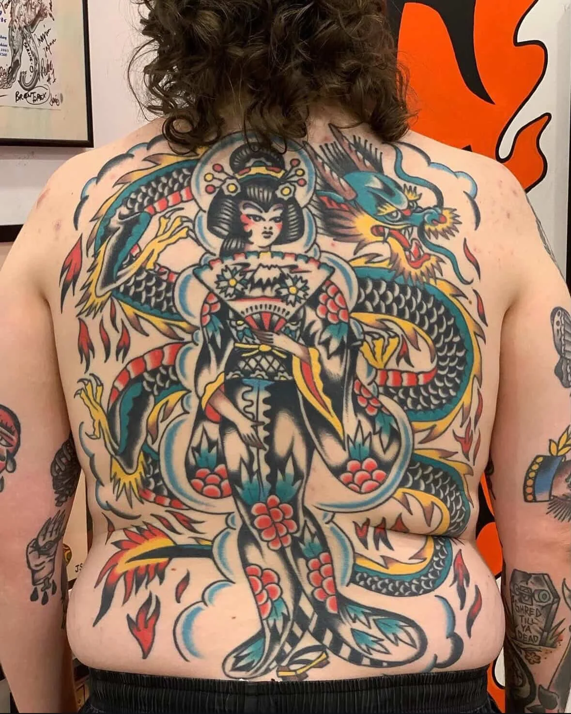 A person with a large colorful tattoo on their chest and stomach, featuring a woman with traditional Japanese clothing and hairstyle, surrounded by a dragon and fiery elements.