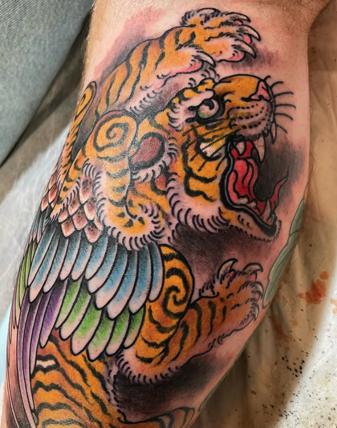 Tattoo of a roaring tiger with colorful wings, featuring orange, black, and pink hues, on a person's arm.