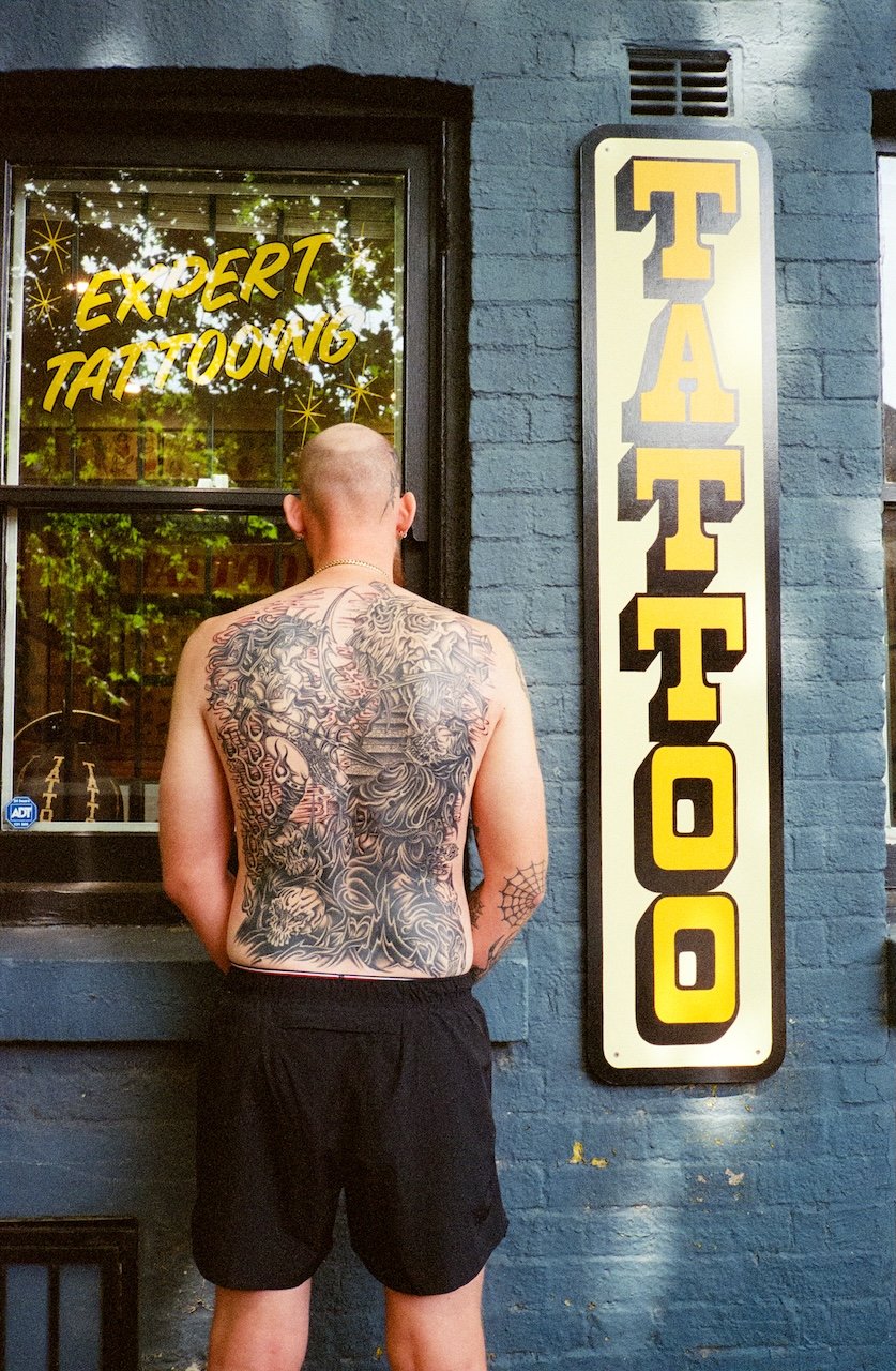 A shirtless man with tattoos on his back standing outside a tattoo shop, which has a vertical sign with the word 'TATTOO' in yellow and black letters, and a window with a sign that says 'Expert Tattooing' in yellow letters. The shop's building is pai