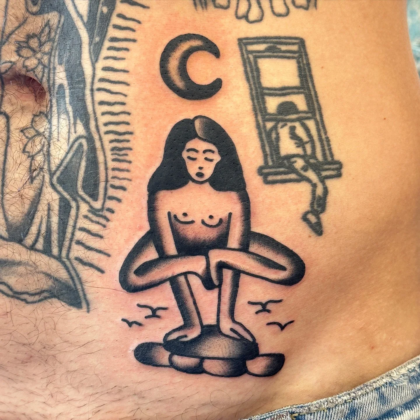 A tattoo of a woman with long hair, sitting with her arms crossed on her knees, with a window and birds in the background, and a crescent moon above her head.