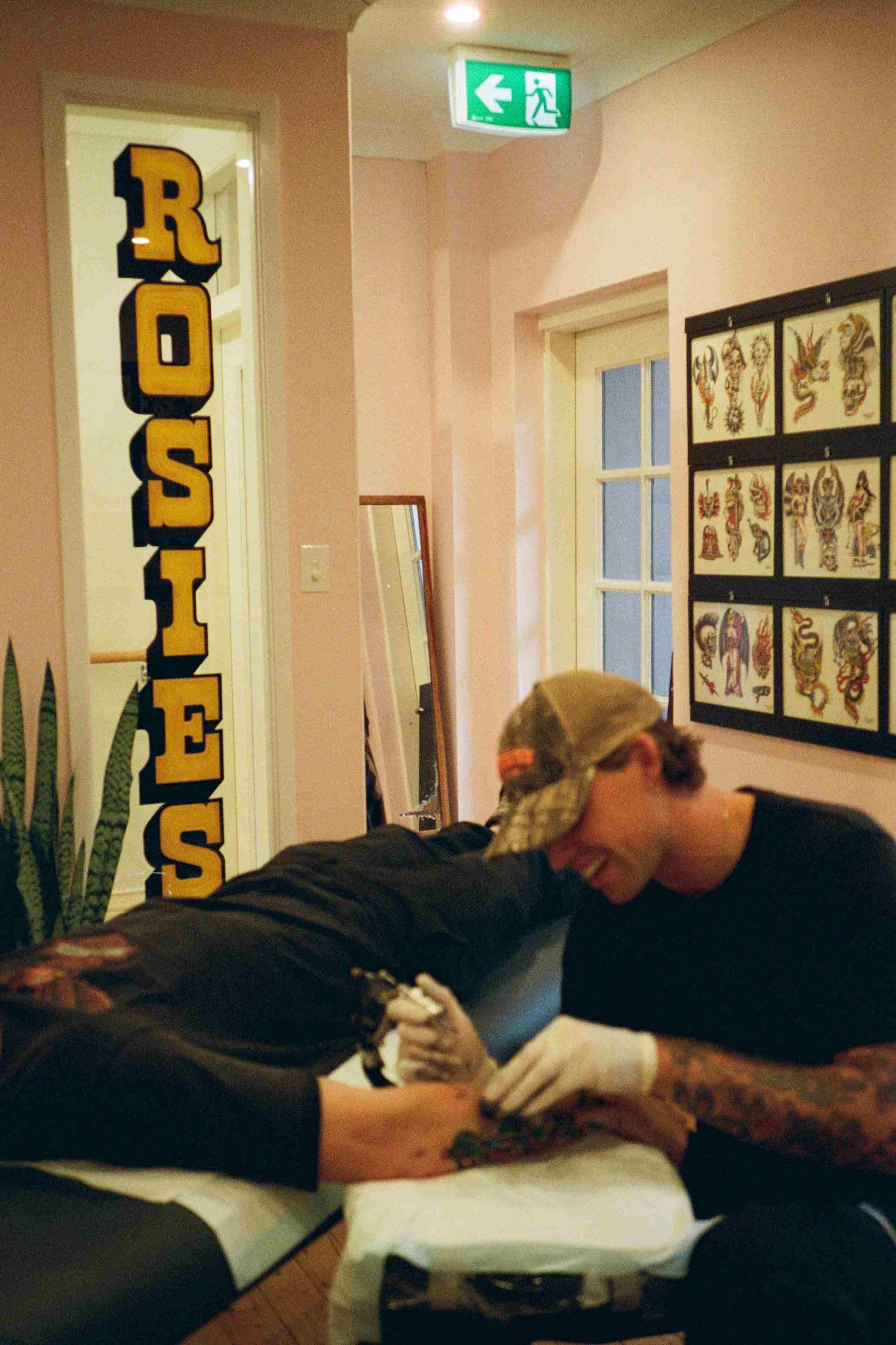 A person with tattoos getting a tattoo on their arm in a tattoo studio, with tattoo flash art on the wall, a mirror, and an emergency exit sign overhead.