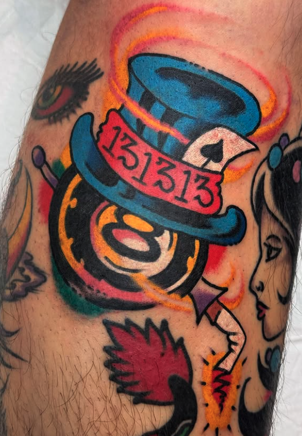 A colorful tattoo on skin featuring a blue top hat with the numbers 13, a playing card with a spade, a clock, an eye with eyelashes, and a woman's face.