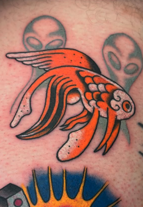 Tattoo of an orange goldfish with flowing fins on skin, with two gray alien faces in the background.