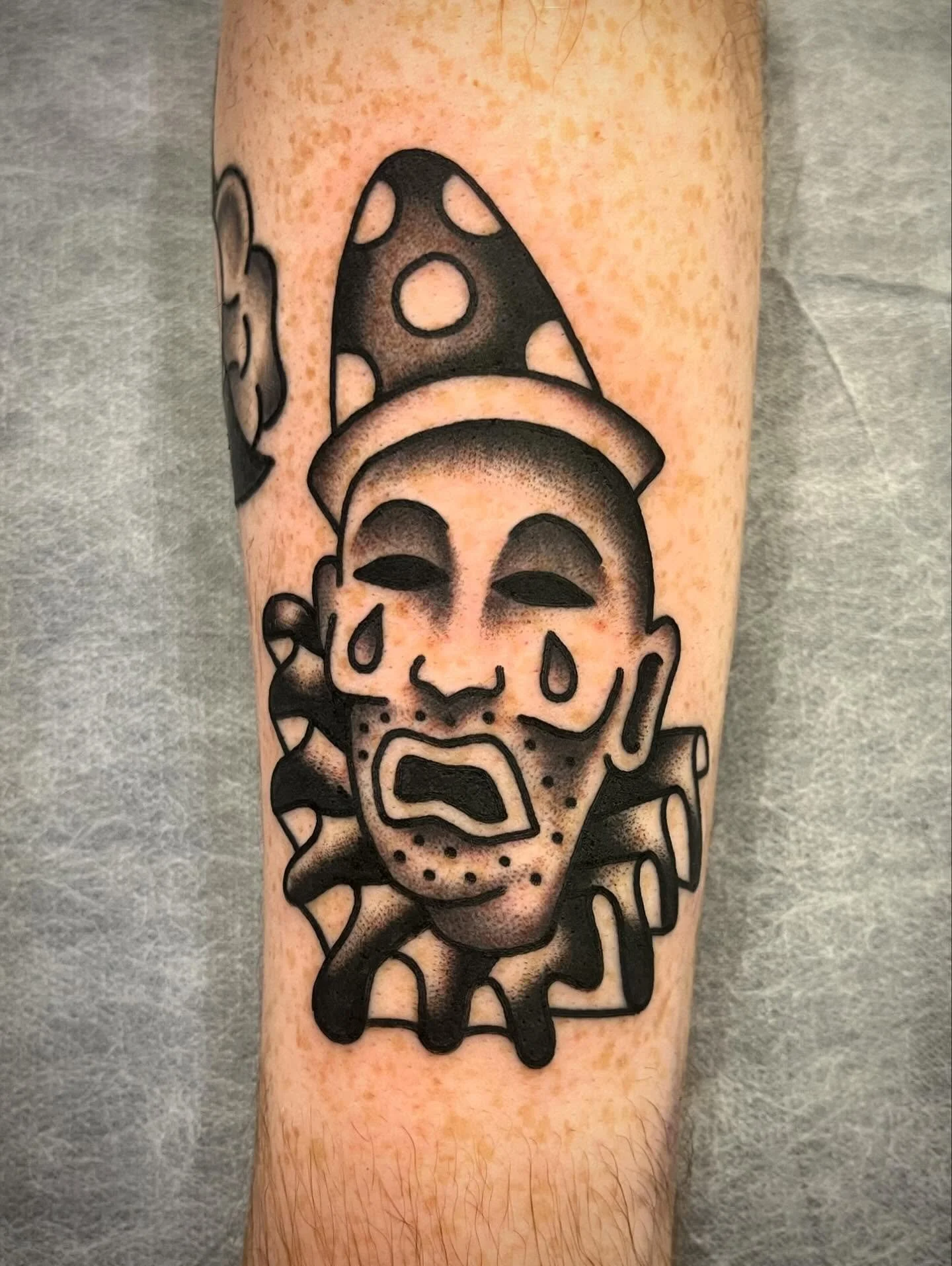 Tattoo of a sad face with tears, a mushroom hat, and a face with a beard, in black ink on a person's arm.
