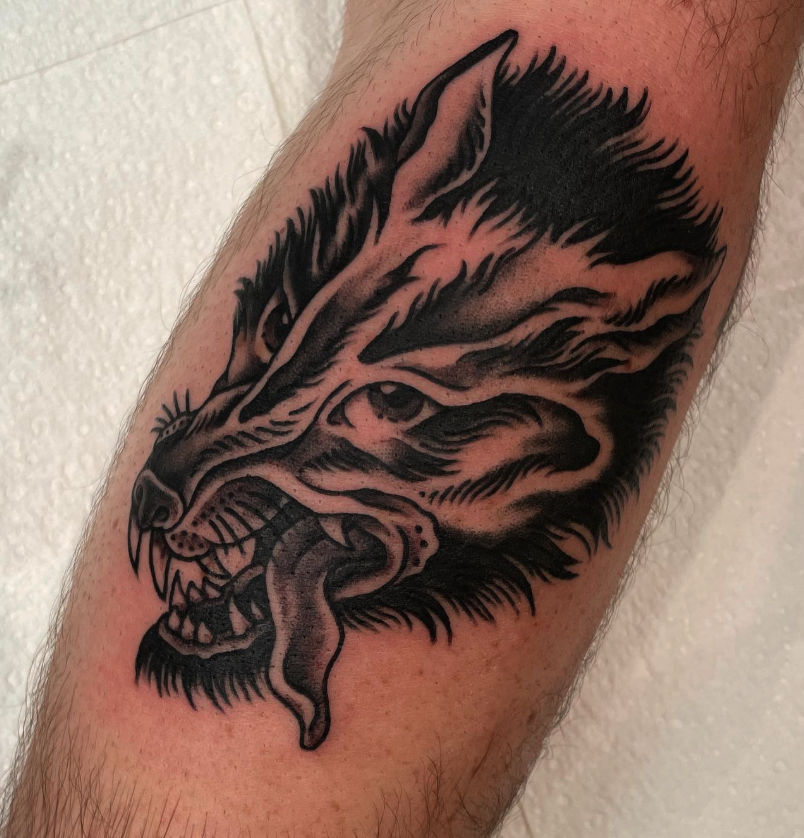 Tattoo of a fierce wolf's head showing sharp teeth and a protruding tongue on a person's arm.