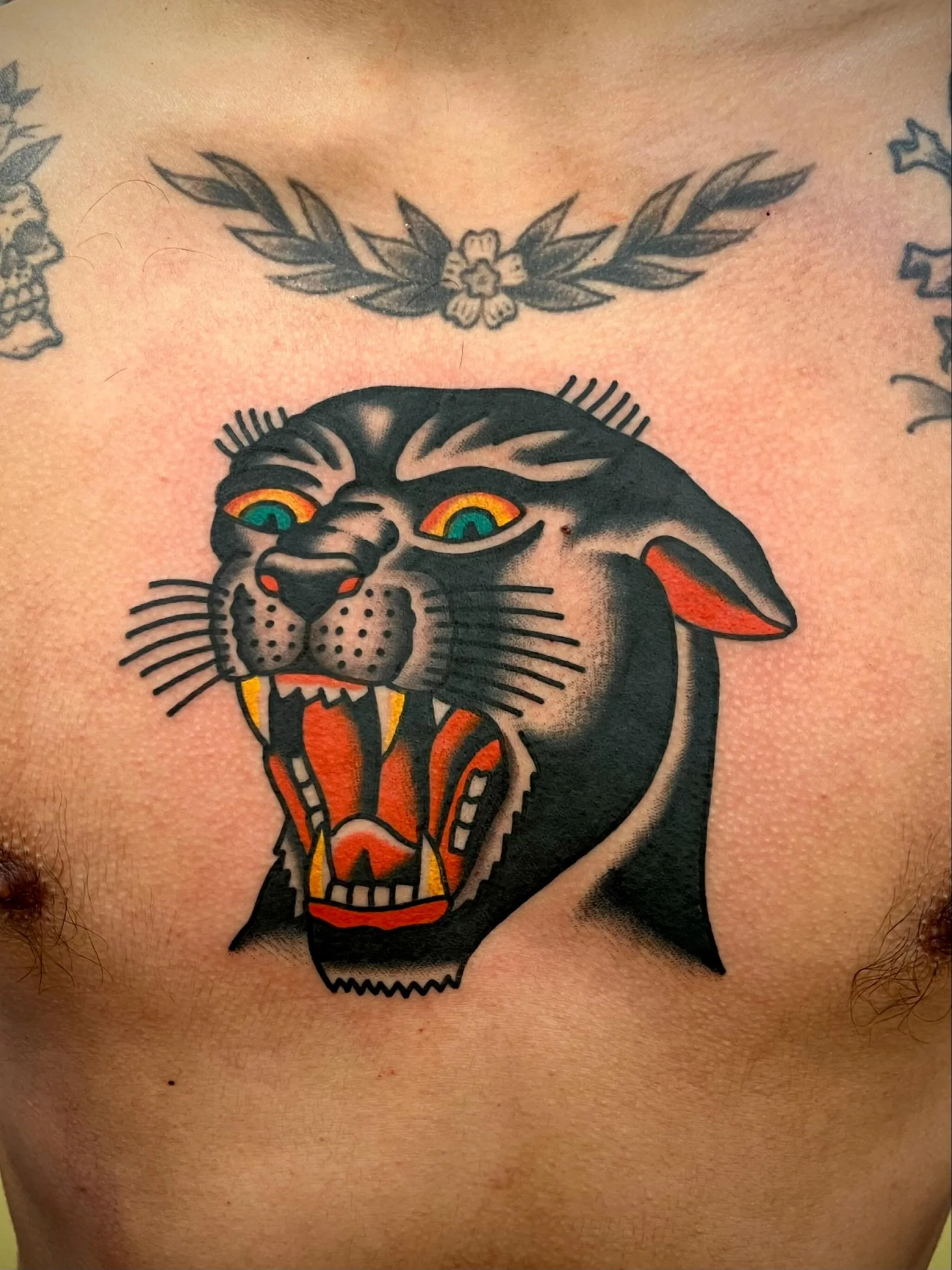 Tattoo of a fierce black panther's head with open mouth, showing teeth and tongue, on a person's chest. Above it, there is a smaller tattoo of a floral wreath with leaves.