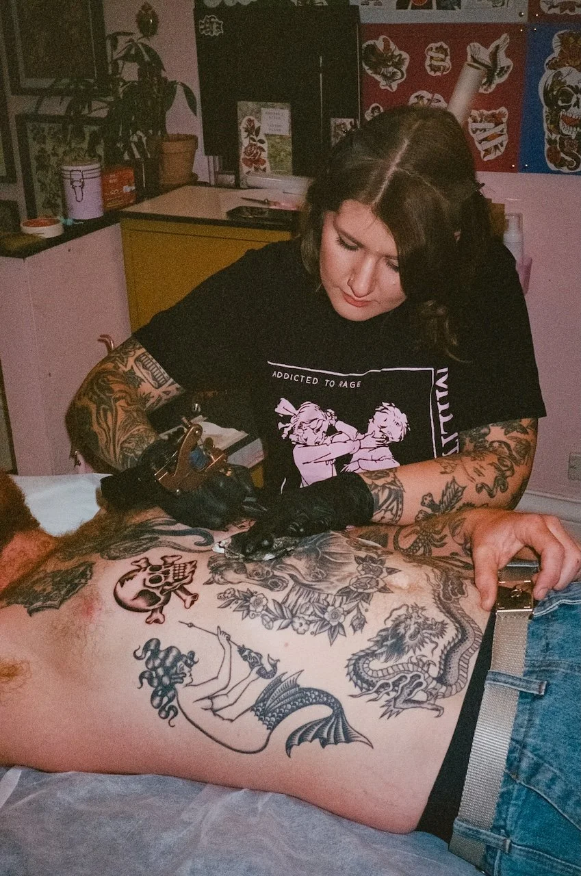 A woman with tattoos on her arms is giving a tattoo to a man laying on a table. The woman is focused on her work, wearing black gloves and holding a tattoo machine. The man has multiple tattoos on his chest and stomach, including a mermaid with a swo