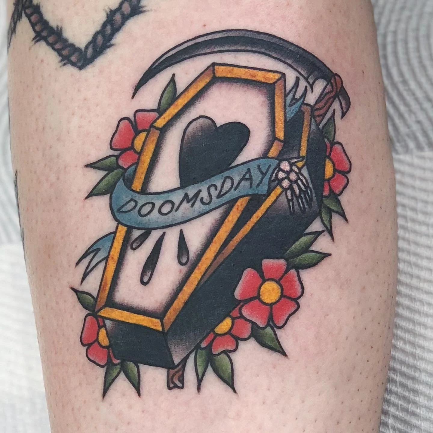 Tattoo of an open coffin with a heart inside, surrounded by red flowers and green leaves, with a banner across it reading 'Doomsday' and black teardrops beneath.