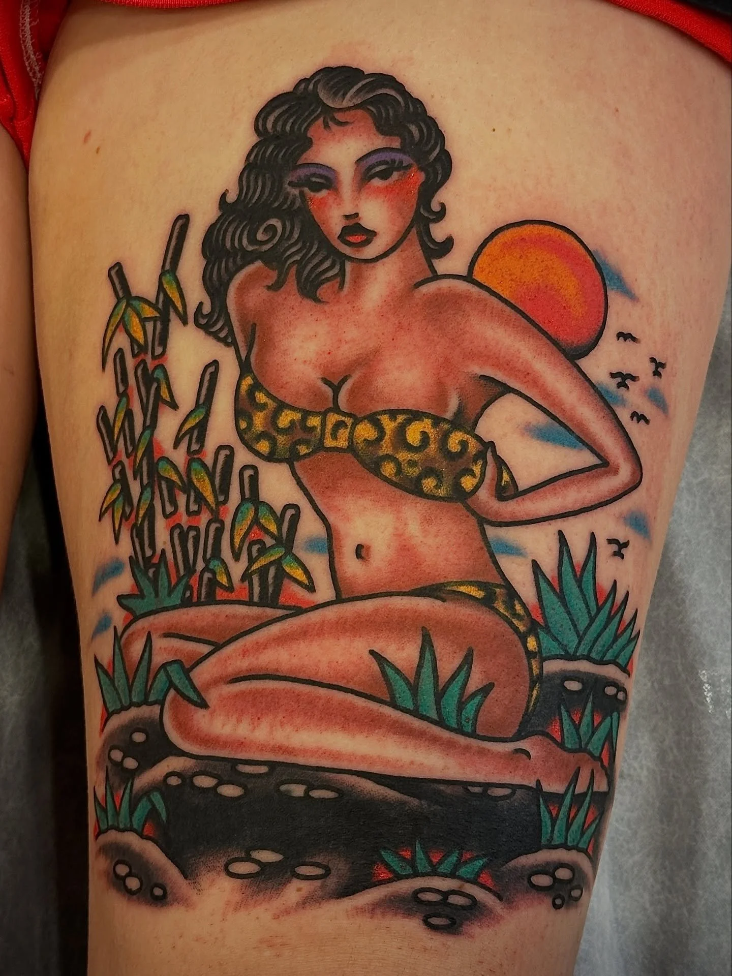 Tattoo of a woman with long, curly black hair, wearing a leopard print top, sitting outdoors with a setting sun, surrounded by green plants and birds.