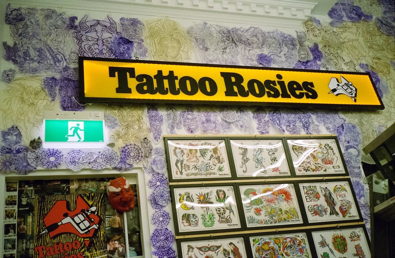 A wall decorated with tattoo artwork and a tattoo shop sign that reads "Tattoo Rosies," with spooky decorations like spider webs and a red devil mask, and an emergency exit sign.