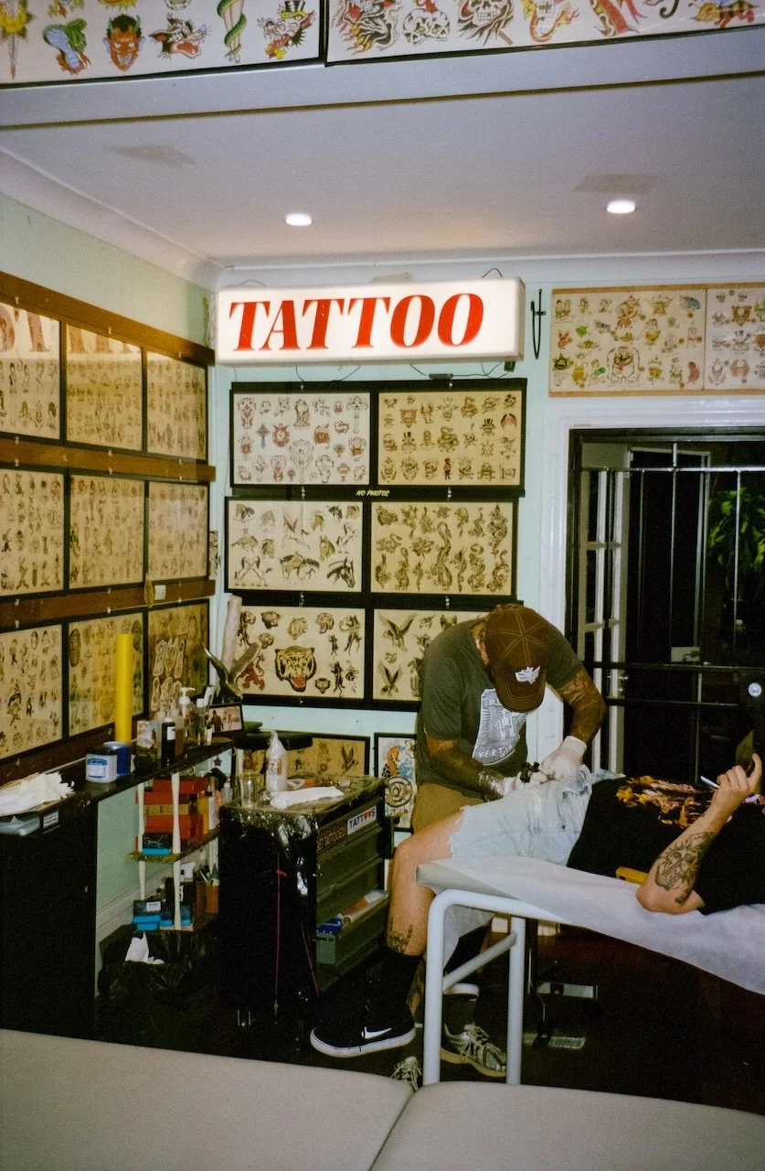 A tattoo studio with tattoo flash artwork on the walls, a lit 'TATTOO' sign, and a person receiving a tattoo from a tattoo artist.