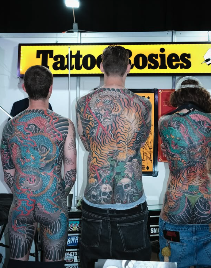 Three people with colorful traditional Japanese-style tattoos on their backs standing at a tattoo booth with a yellow sign reading "Tattoo Rosies."