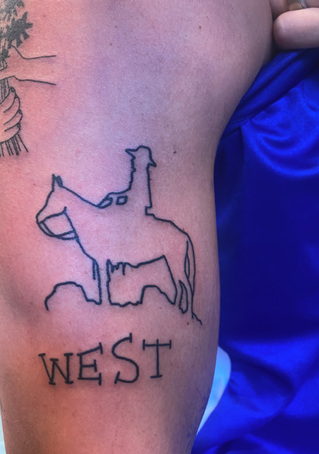 Tattoo of a dog with a saddle and the word 'WEST' beneath it on a person's thigh.