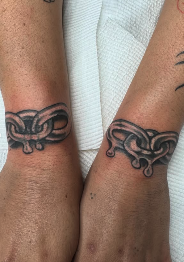 Two forearms with matching black and gray tattoo designs of a stylized snake intertwined with its head and tail near the wrists.