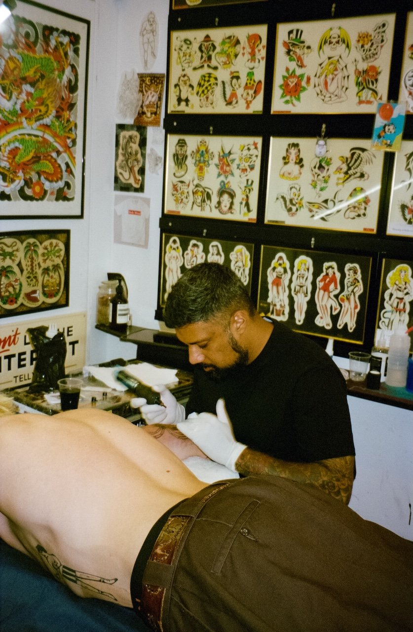 A tattoo artist working on a person's side in a tattoo studio decorated with flash art on the walls, including traditional-style tattoos of women, dragons, and other designs.