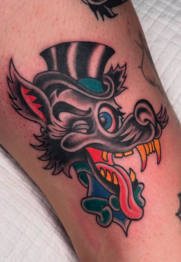 Colorful tattoo of a mischievous or angry wolf with exaggerated features, wearing a striped hat, with sharp teeth, a long tongue, and detailed fur on the arm.