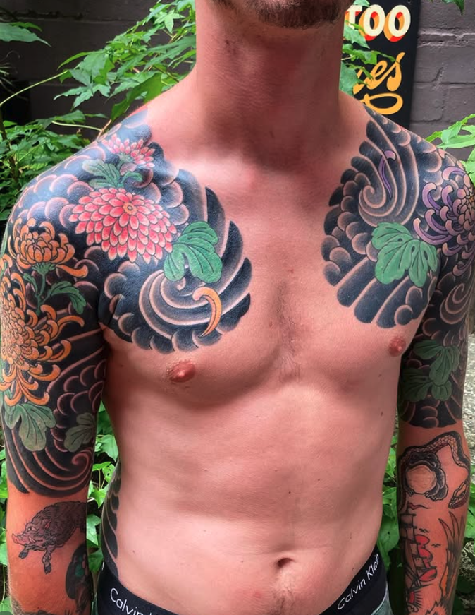 Close-up of a shirtless man with extensive colorful tattoos on his chest, shoulders, and arms, including flowers, leaves, and a snake, with a background of green foliage and a black brick wall.
