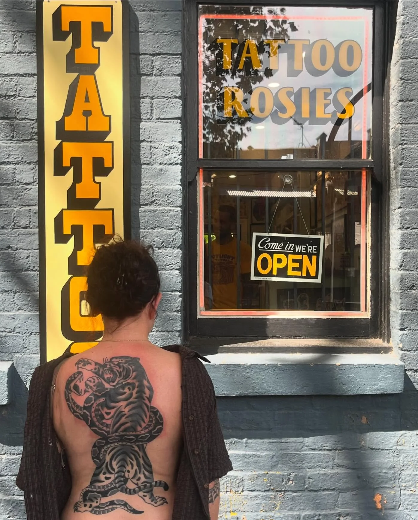 A person with a large black tattoo of a tiger on their back, standing outside a tattoo shop. The shop has a yellow and black vertical sign that reads 'TATTOO' and a window with signs that say 'Tattoo Rosies' and 'Come in we're open' in black and yellow.