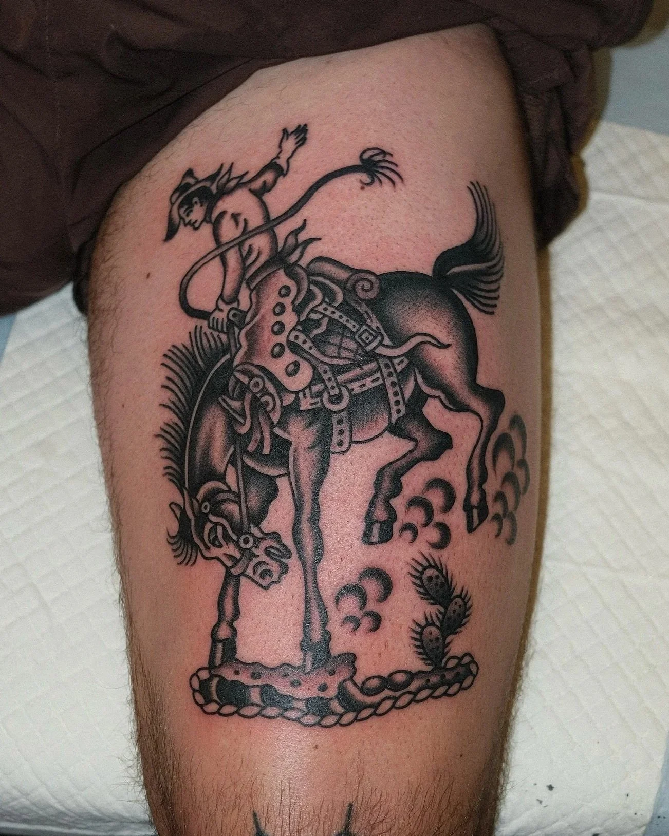 Tattoo of a cowboy riding a bucking horse, with smoke or clouds and a small cactus nearby.