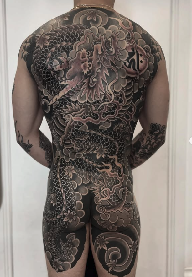 Back and legs of a person with extensive black and gray traditional Japanese-style tattoos featuring a dragon, cherry blossoms, and cloud motifs.
