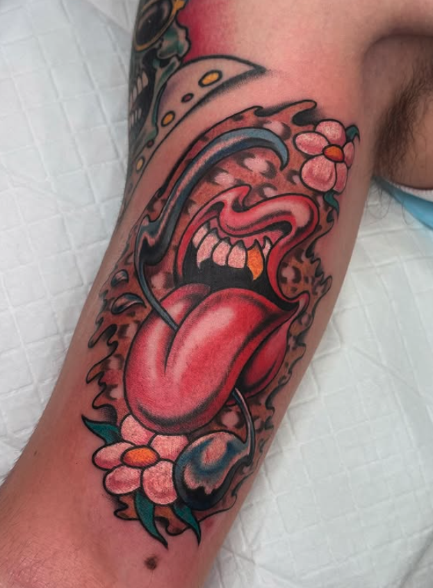 Colorful tattoo on a person's arm depicting a stylized, cartoonish face with a large open mouth, tongue sticking out, a prominent nose, orbits, surrounded by pink flowers and leaves.