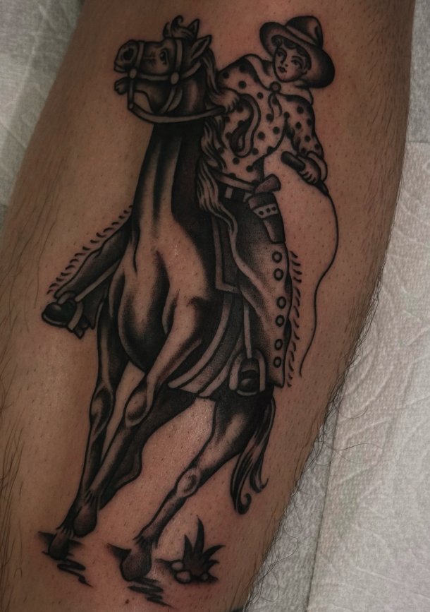 Tattoo of a cowboy riding a horse, holding a whip, with a hat and scarf, in black ink style.