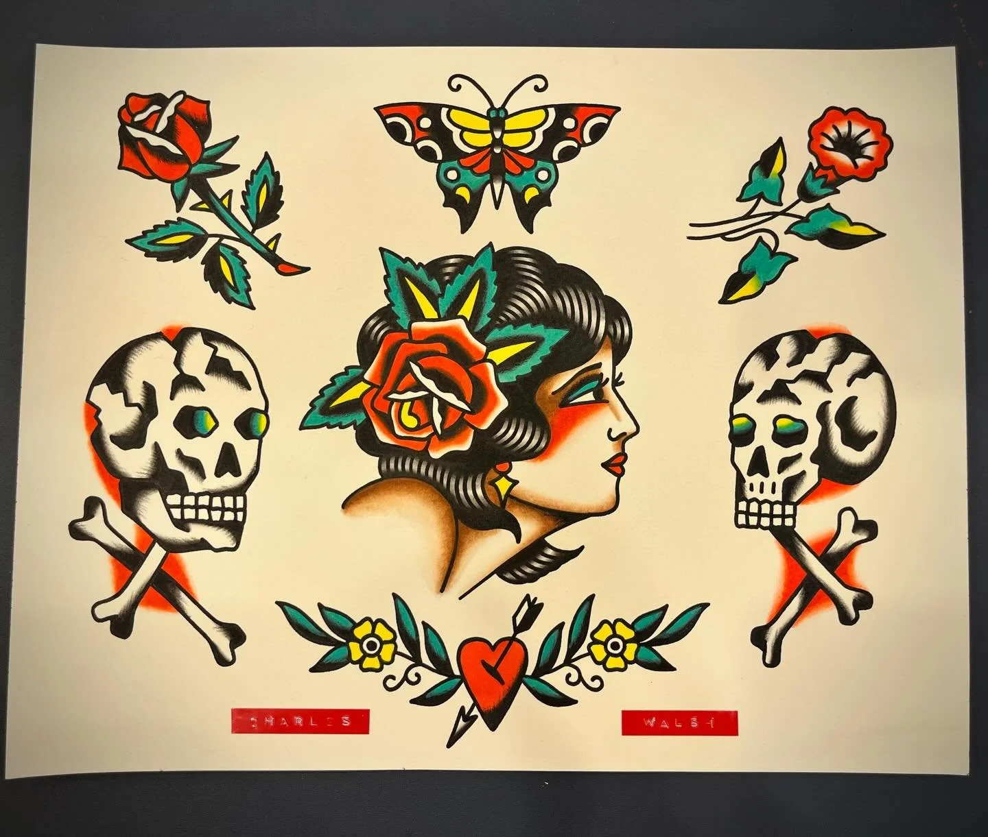 Tattoo-style artwork featuring a woman with dark hair and colorful flowers in her hair, surrounded by two skulls with crossed bones, a butterfly above her, and roses and leaves. There are red labels with the names 'Charles' and 'Walsi' at the bottom.