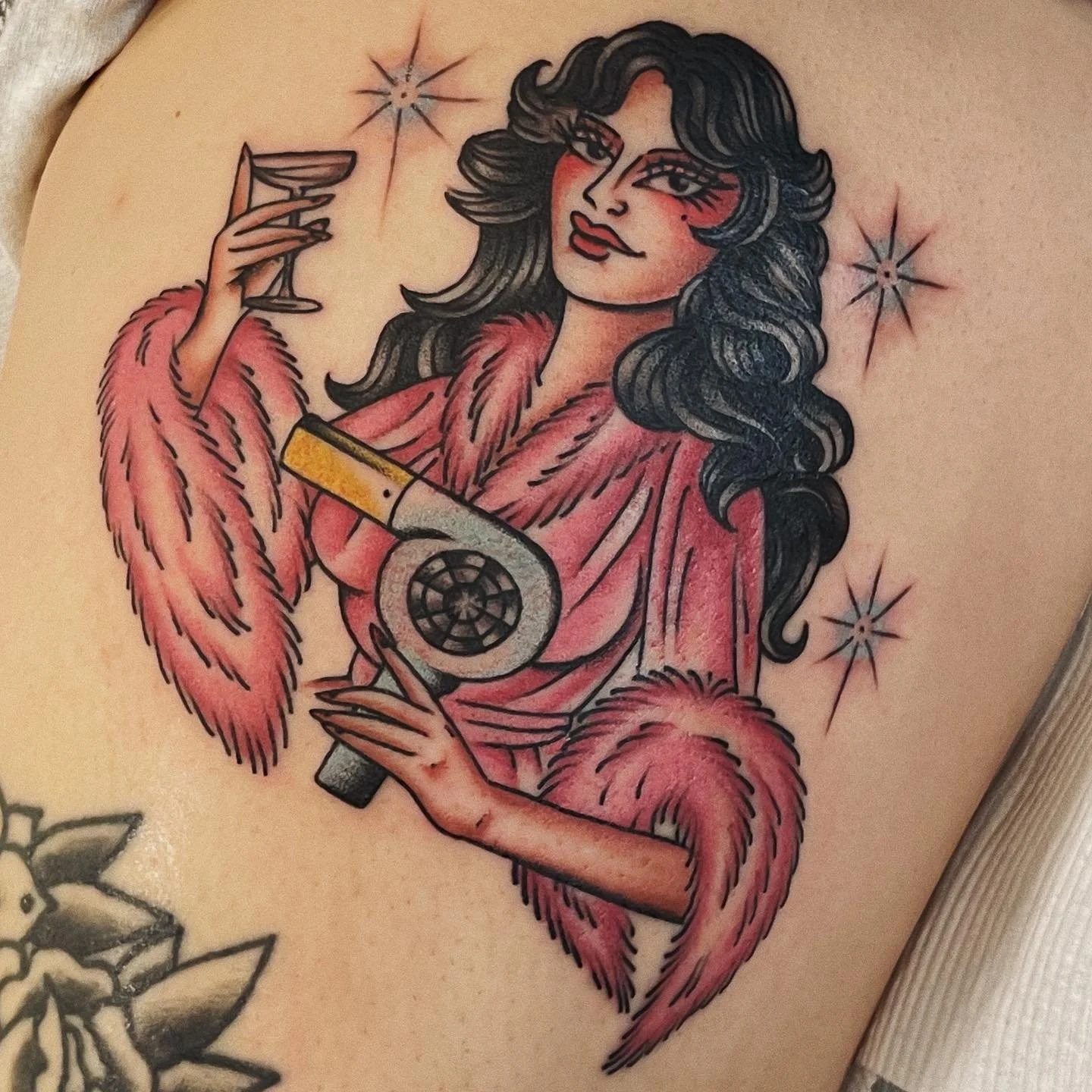 A tattoo of a woman with long dark hair, wearing a pink fur coat, holding a vintage hairdryer in one hand and a glass of wine in the other, with stars around her head.