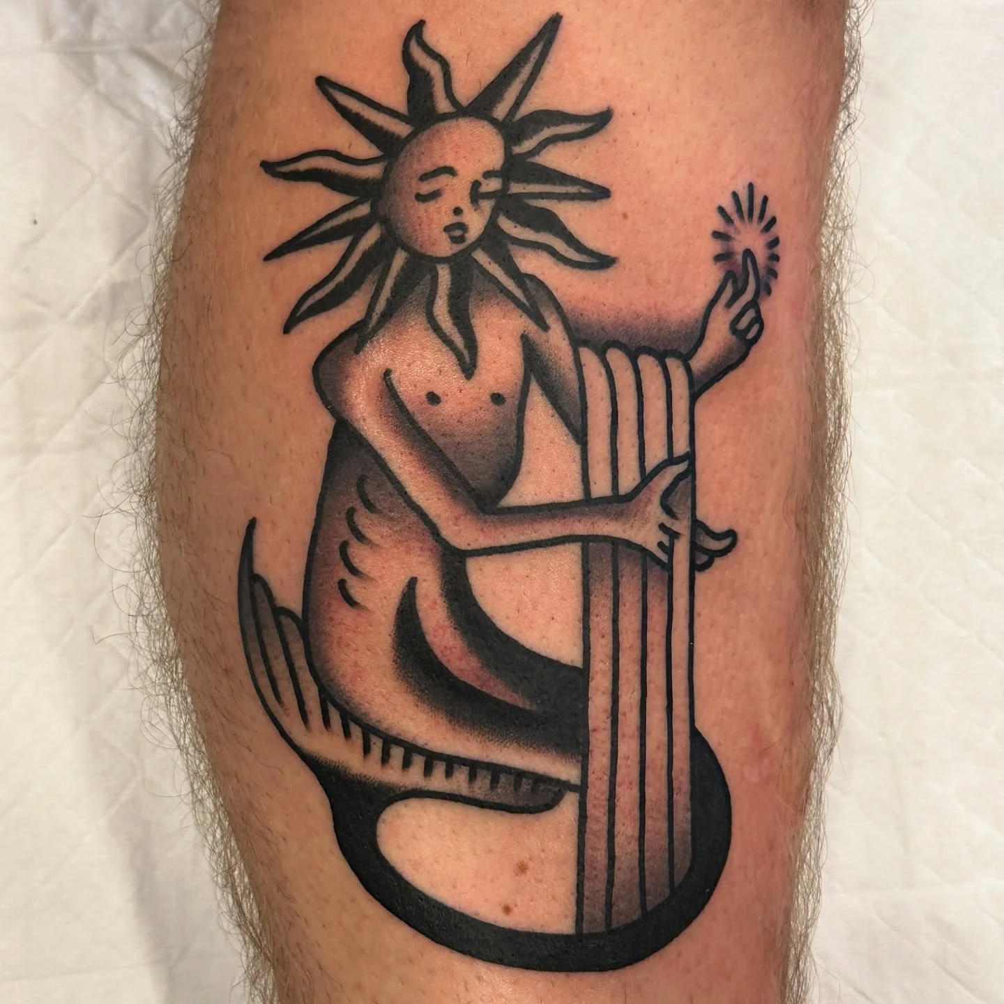 Tattoo of a mermaid with a sun as a head, holding a small sun in one hand, with a black and gray shading style.