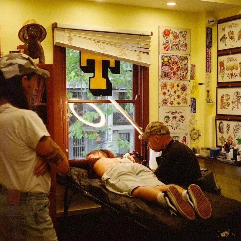 Tattoo artist tattooing person's back while person lies on tattooing bed at tattoo parlor with tattoo designs on walls.