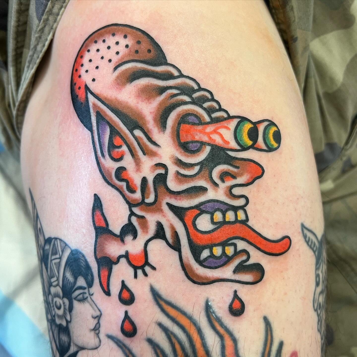 Colorful tattoo of a cartoonish, monstrous face with exaggerated features, big eyes, and a long tongue, with dripping red ink and detailed shading.