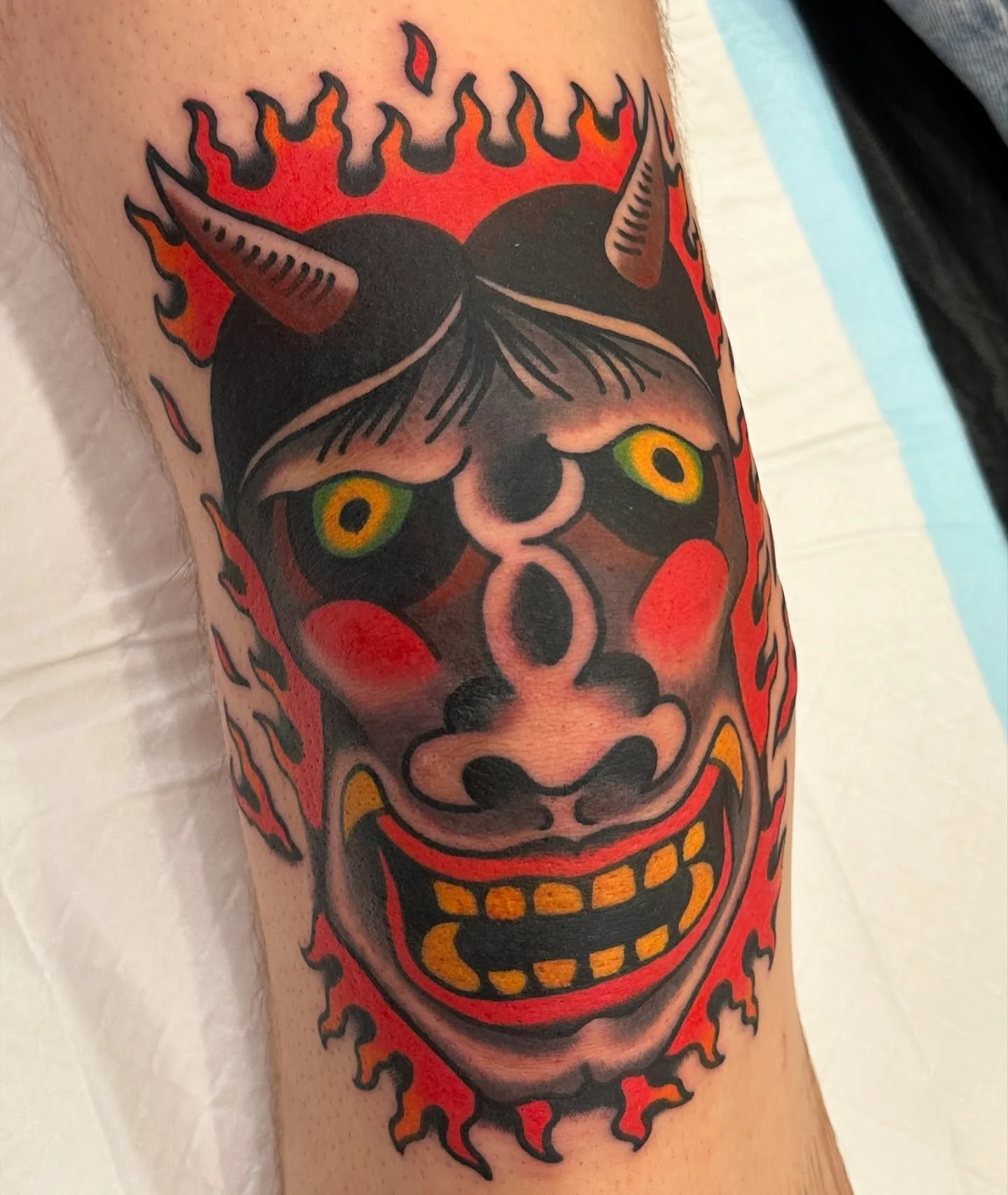 Traditional tattoo of an Oni mask with flames around it, featuring horns, piercing yellow eyes, and a wide grin with yellow teeth.