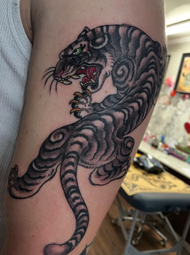 Tattoo of a fierce tiger with open mouth, sharp teeth, and green eyes, wrapped around the upper arm.