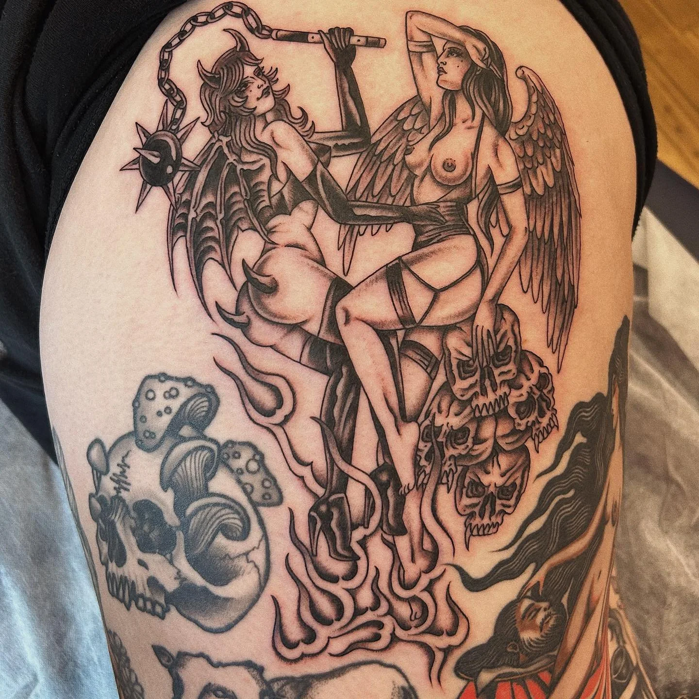 A detailed tattoo featuring two winged women, one with devilish features and the other with angelic features, engaging in an intimate pose. The tattoo also includes skulls, mushrooms, and flames.