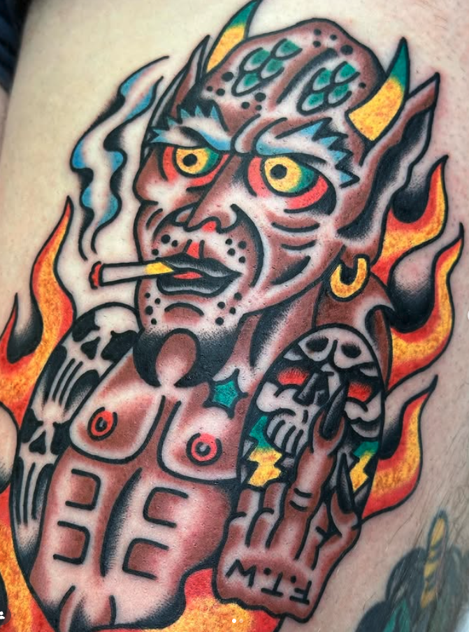 Tattoo of a devil-like figure with yellow eyes, smoking a cigarette, with flames and skulls surrounding it.