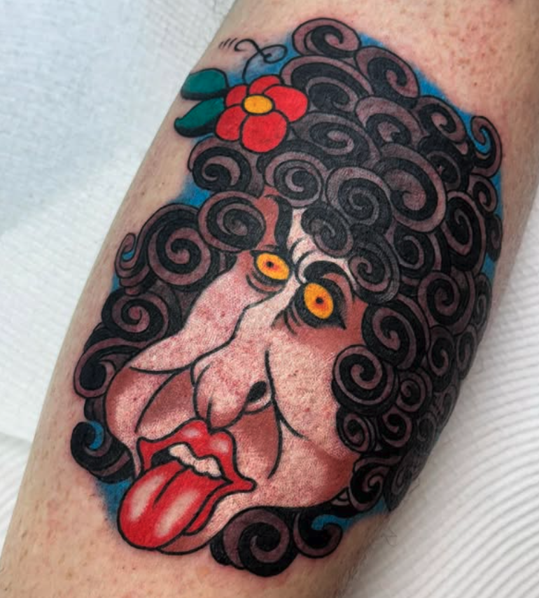 Tattoo of a distorted face with curly black hair, yellow eyes, red tongue sticking out, and a red flower with green leaves above the head.