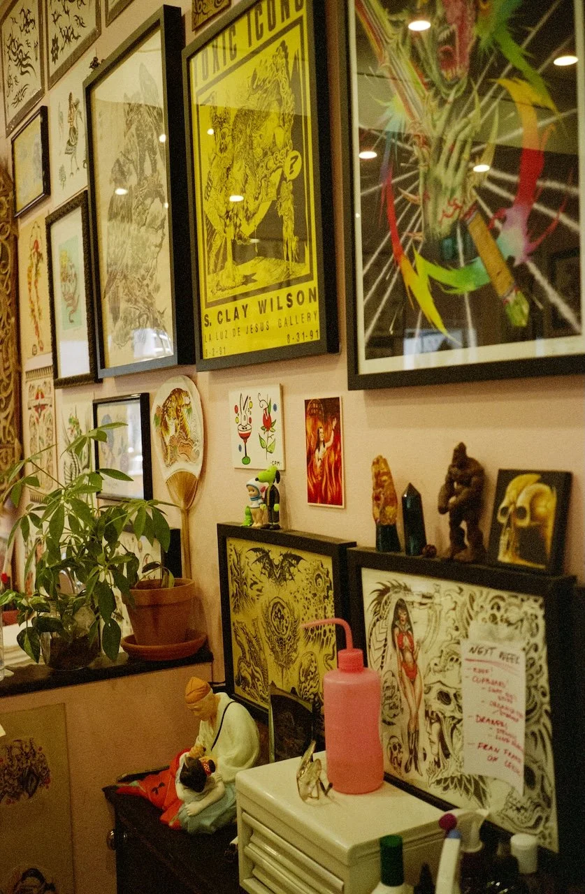 Wall decorated with framed artwork and posters, including a yellow S. Clay Wilson poster, a colorful dragon illustration, and various smaller pieces. A potted plant is on a shelf, along with figurines, sculptures, and a pink water bottle. There is a 
